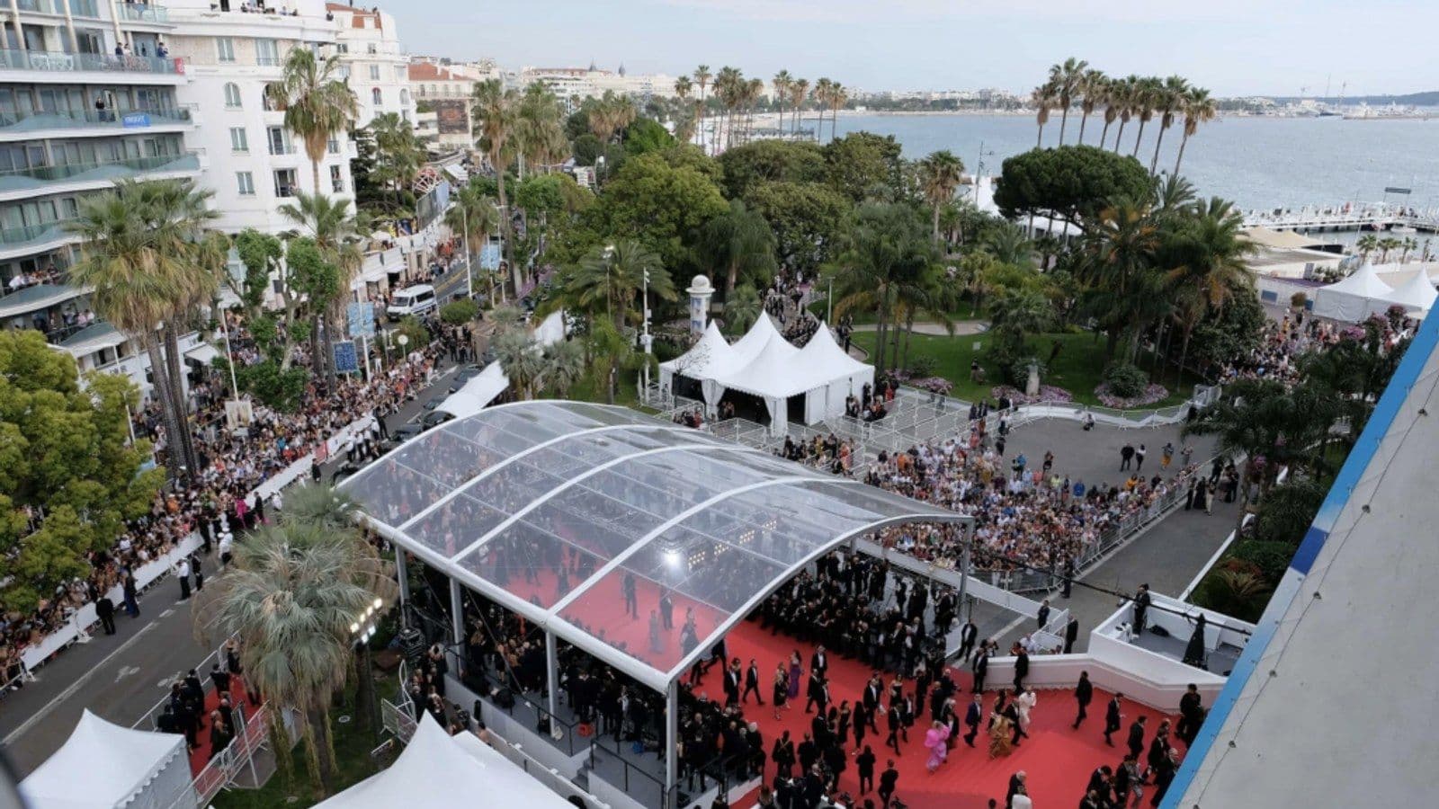 Vista aérea de la alfombra roja de Cannes bajo su marquesina acristalada, con invitados llegando, público tras las vallas a lo largo de la calle y el mar al fondo.