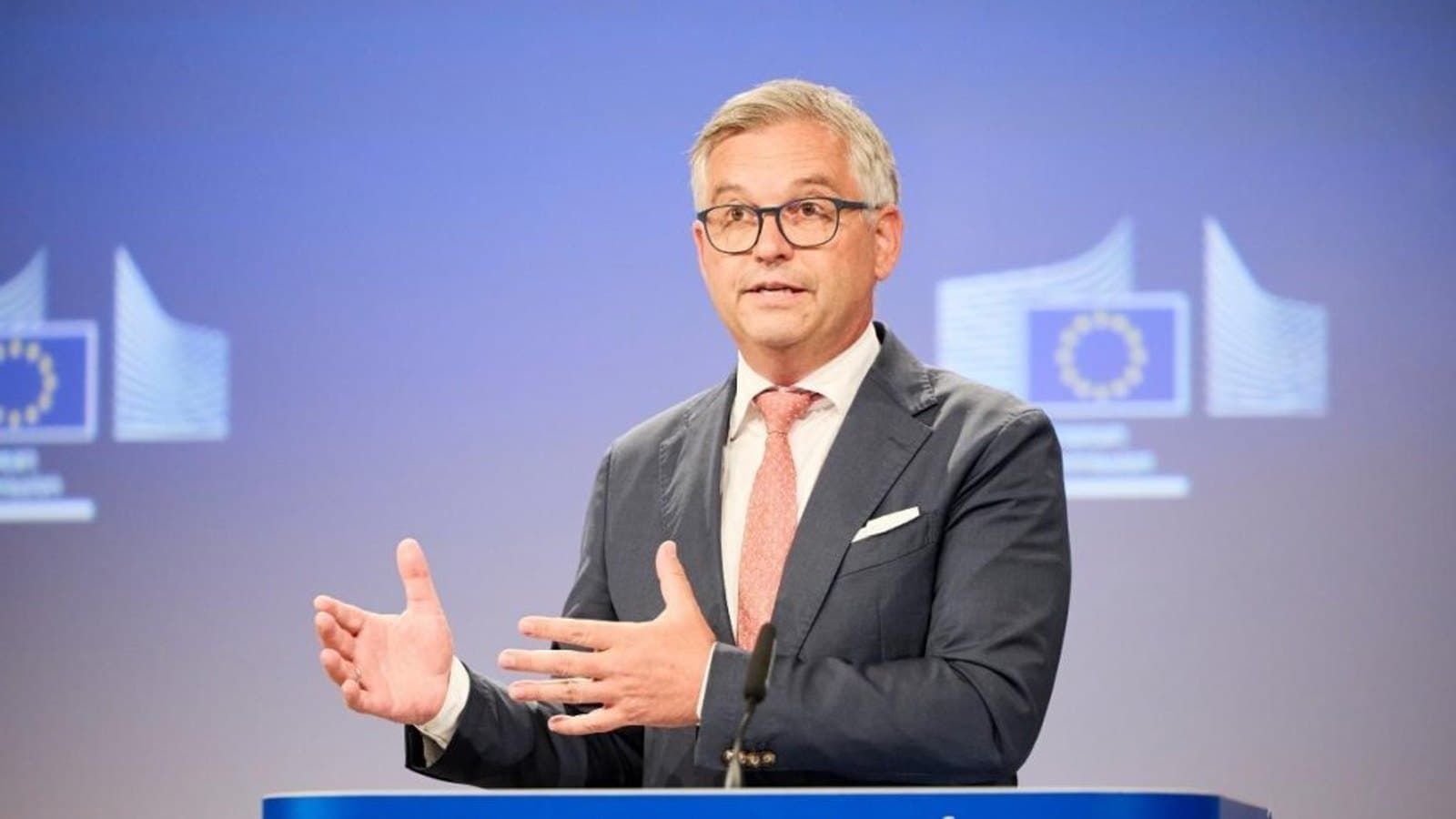 Un hombre con traje oscuro y corbata rosa gesticula con ambas manos mientras habla en un atril con el rótulo Comisión Europea, con emblemas de la UE en la pared detrás.