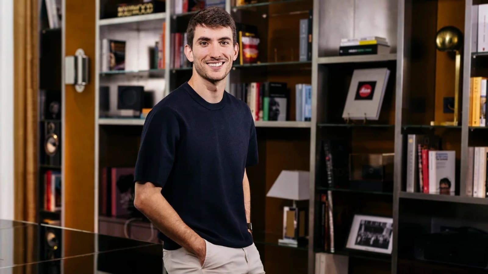 Pablo Palafox sonríe con un jersey oscuro de cuello redondo, de pie ante una estantería con libros y objetos.