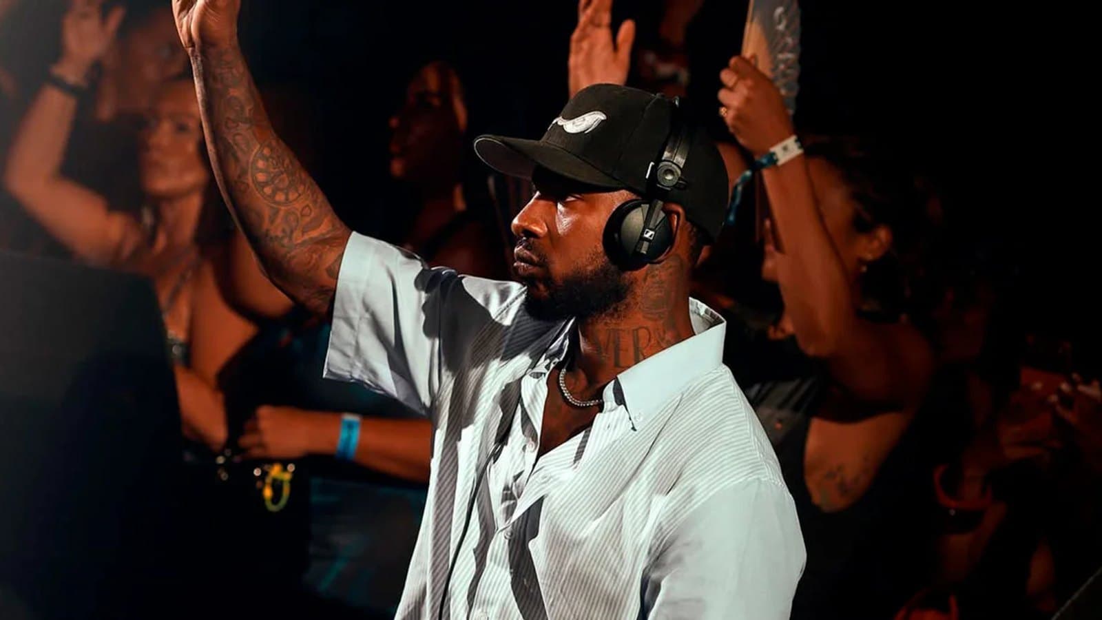 Skepta pinchando en el escenario con gorra y auriculares, un brazo levantado, con el público detrás con las manos en alto.