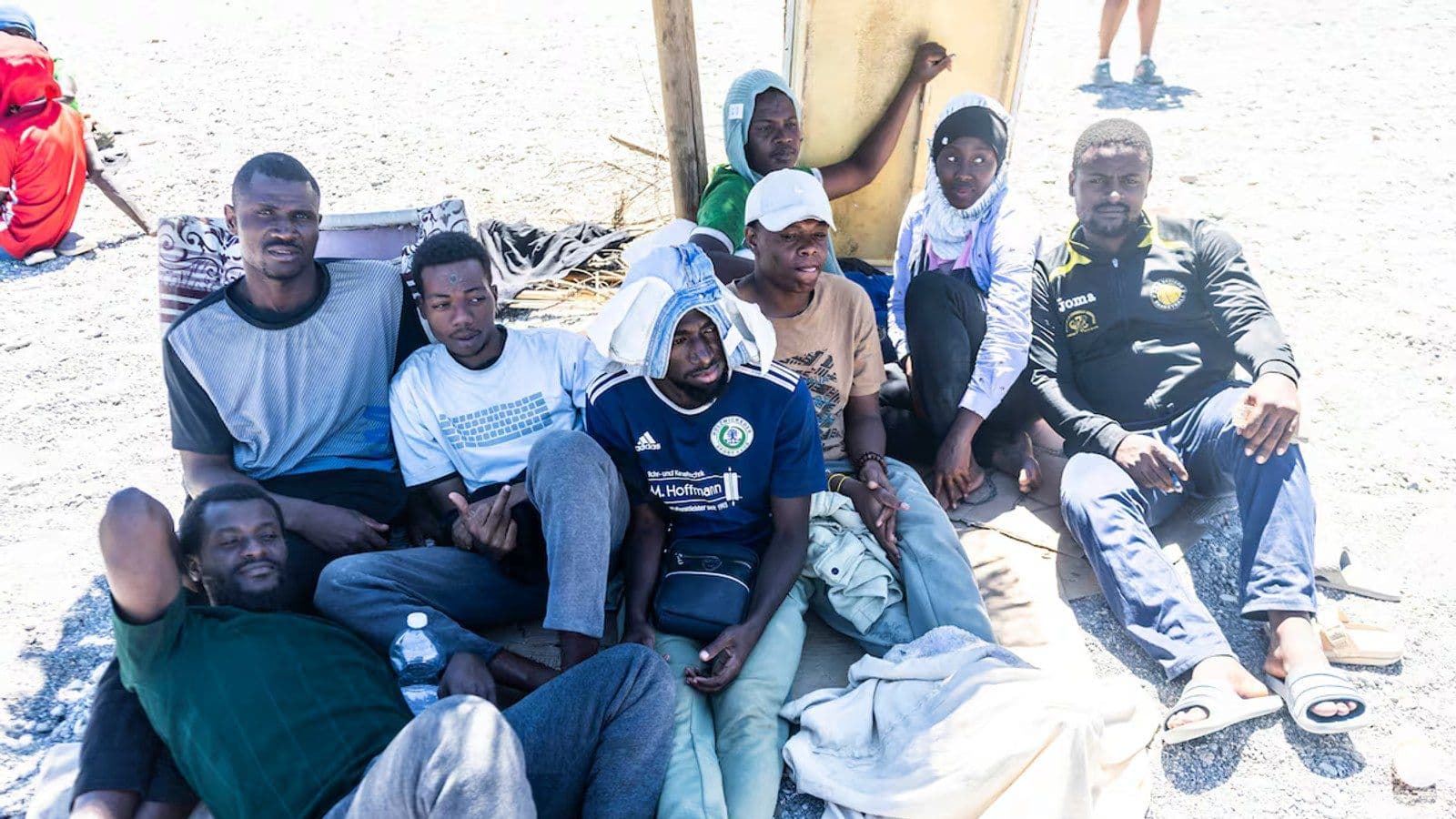 Migrantes reunidos en una pequeña franja de arena en Ceuta, resguardándose del sol bajo mantas y refugios improvisados.