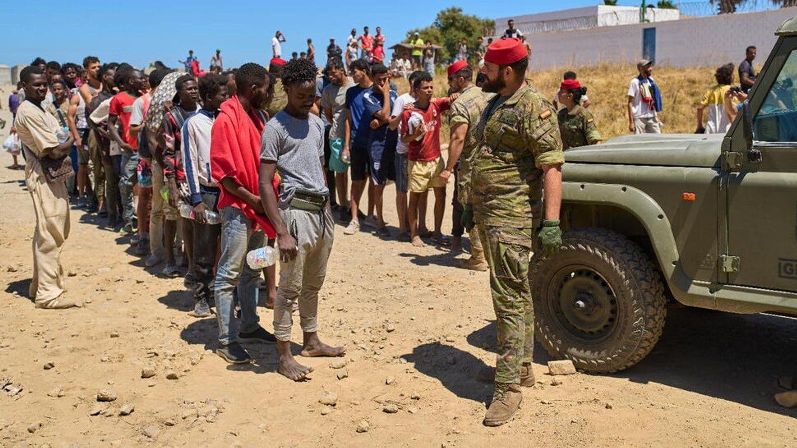 Una fila de jóvenes, la mayoría con poco más que botellas de agua, espera en un terreno seco junto a un militar español de camuflaje y un vehículo militar, con más personas reunidas en una ladera al fondo.